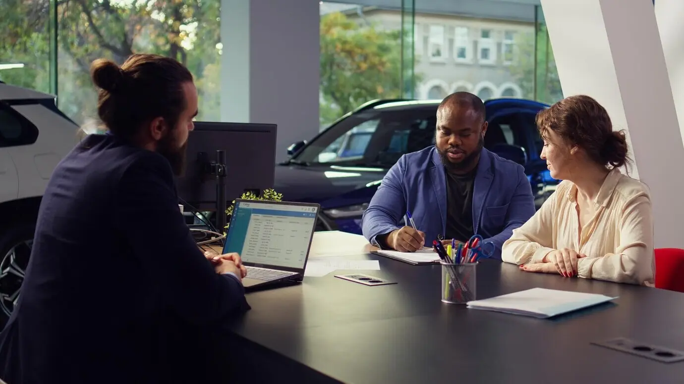 Продавец автосалона сидит за столом с клиентами и передаёт им контракт для подписи.