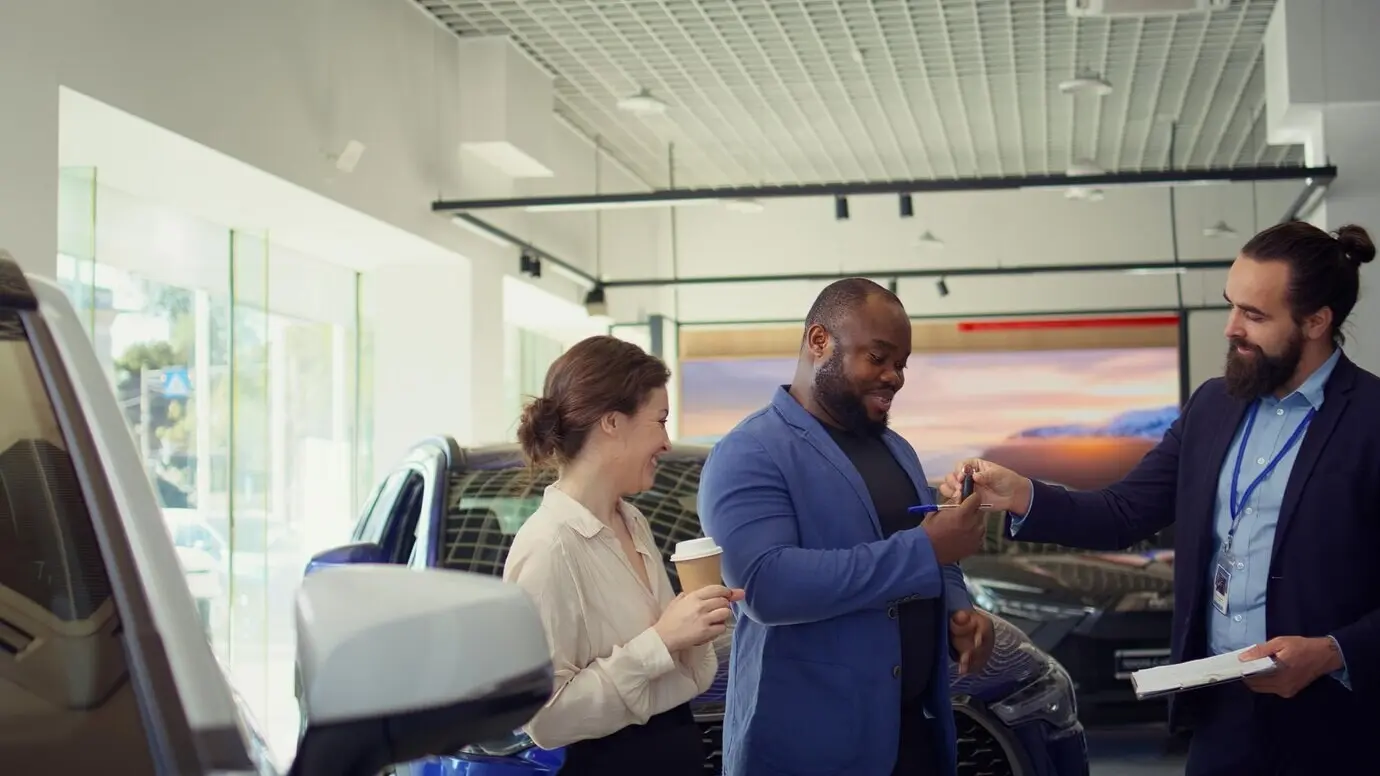 Продавец автомобилей передает клиентам договор купли-продажи для подписи после того, как предложил им ключи от автомобиля.